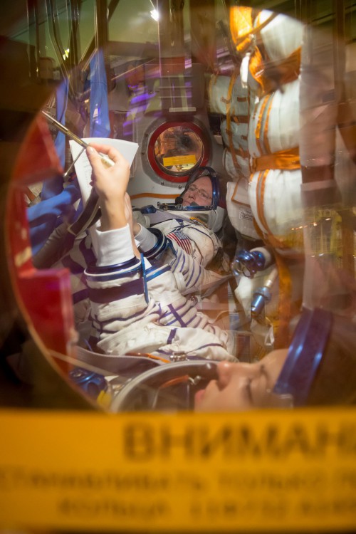 The Soyuz TMA-14M crew participate in a "fit check" within the cramped confines of the "real" descent module of their spacecraft. Barry "Butch" Wilmore is visible in the background, with Yelena Serova in the foreground and Aleksandr Samokutyayev partially obscured in the center seat. Photo Credit: NASA