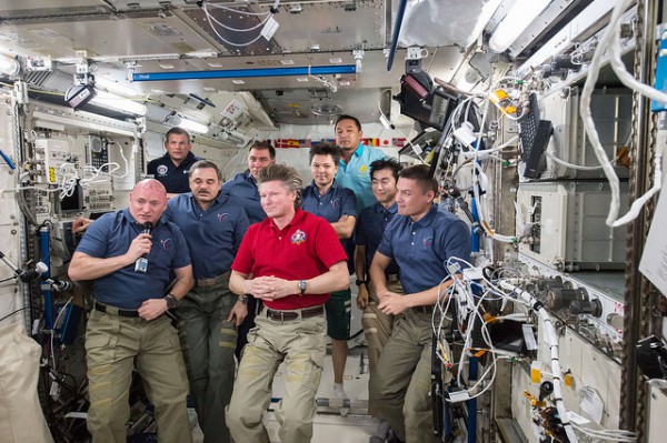 Expedition 45 Commander Scott Kelly (with microphone, front left) accepts command of the space station from outgoing Expedition 44 Commander Gennadi Padalka (in red shirt), as their crewmates look on. Photo Credit: NASA