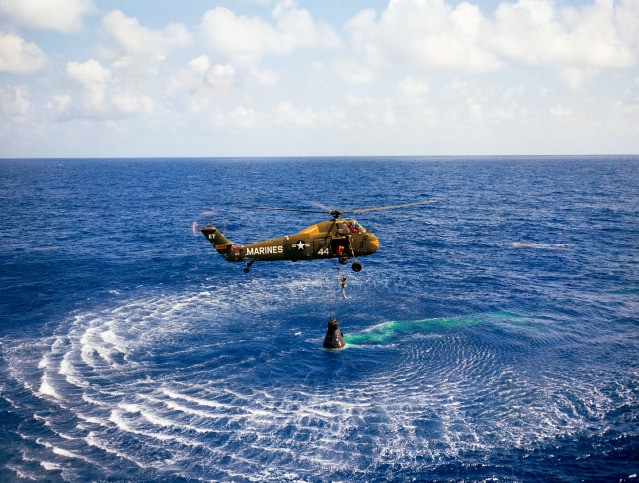 Alan Shepard is hoisted aboard the helicopter, deftly piloted by Wayne Koons, after completing his 15-minute suborbital flight. The patch of fluorescent green marker dye in the water around Freedom 7 is particularly obvious. Photo Credit: NASA