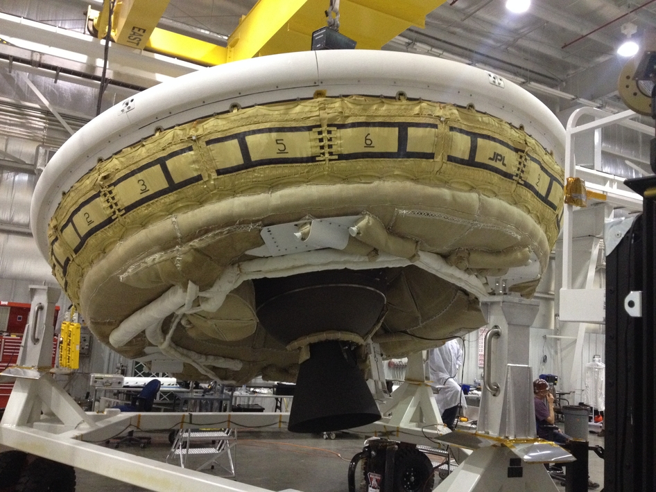The saucer-shaped LDSD test vehicle holding equipment for landing large payloads on Mars is shown in the Missile Assembly Building at the US Navy's Pacific Missile Range Facility in Kaua‘i, Hawaii.  The vehicle, part of the Low Density Supersonic Decelerator project, will test an inflatable decelerator and a parachute at high altitudes and speeds over the Pacific Missile Range this summer.  A balloon will lift the vehicle to high altitudes, where a rocket will take it even higher to the top of the stratosphere at several times the speed of sound. This image was taken during a "hang-angle" measurement, in which engineers set the vehicle's rocket motor to the appropriate angle for the high-altitude test. The nozzle and the lower half of the Star-48 solid rocket motor are the dark objects seen in the middle of the image below the saucer. Photo Credit: NASA / JPL-Caltech