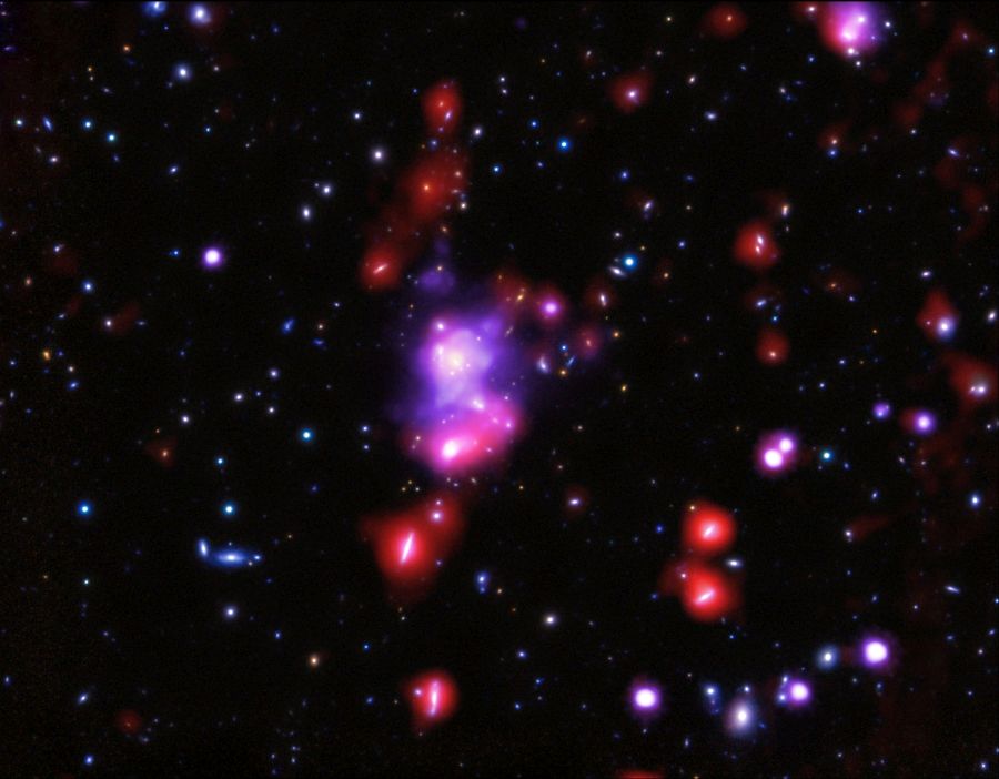 A composite, multi-wavelength image of the young galaxy cluster XDCP J0044.0-2033, also known as the Gioiello Cluster, which is located 9.6 billion light-years away. In the image, X-ray wavelengths appear purple, infrared appear as large red halos around some galaxies, and visible light appears as are red, green, and blue. Image Credit:  X-ray: NASA/CXC/INAF/P.Tozzi, et al; Optical: NAOJ/Subaru and ESO/VLT; Infrared: ESA/Herschel