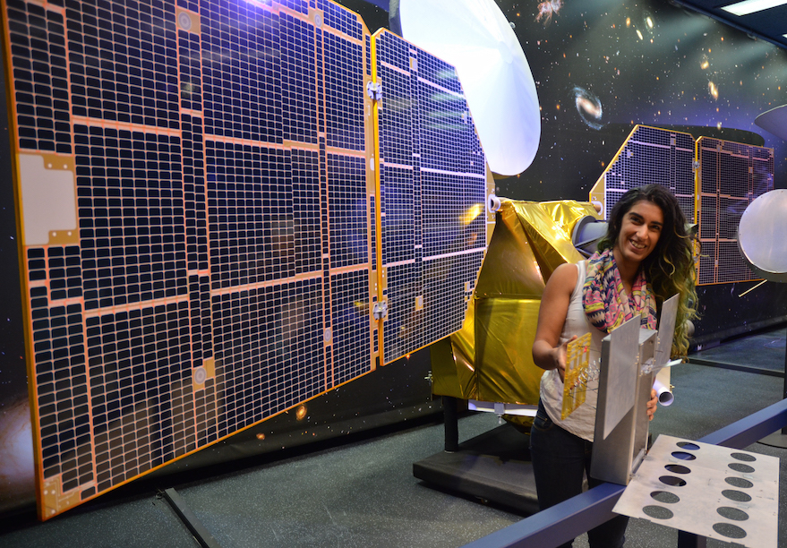 A full-scale mockup of MarCO, held by Farah Alibay, a systems engineer for the technology demonstration. A one-half-scale model of the Mars Reconnaissance Orbiter is behind her. Photo Credit: NASA/JPL-Caltech