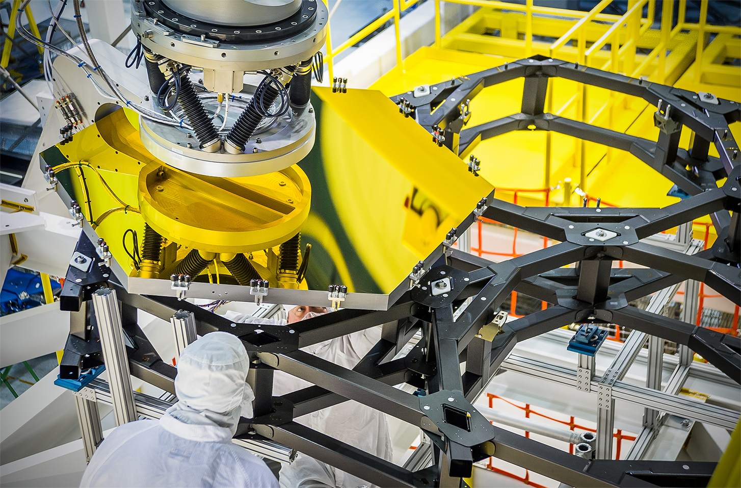 A robotic arm lifts and lowers a golden James Webb Space Telescope flight spare primary mirror segment onto a test piece of backplane at NASA's Goddard Space Flight Center in Greenbelt, Md. In this image the JWST team practices the positioning that will be done on the actual telescope in the cleanroom. Dave Sime, an assembly crew chief, inspects the mirror placement from the underside of the backplane. Photo Credit: NASA/Chris Gunn